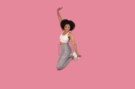 Foto de Enérgica joven afroamericana con el pelo rizado saltando alegremente en el aire, vestida con sujetador deportivo blanco y polainas grises, aislada sobre fondo rosa - Imagen libre de derechos