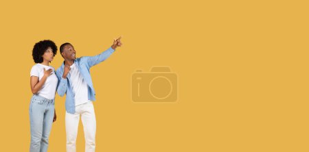 Téléchargez les photos : Enthousiaste jeune couple noir souriant avec des sourires lumineux pointant vers l'extérieur, s'engageant avec quelque chose d'intéressant hors caméra sur un fond jaune moutarde spacieux, studio - en image libre de droit