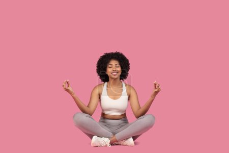 Téléchargez les photos : Souriante jeune femme afro-américaine profite d'un moment paisible en position lotus, pratiquant la pleine conscience avec les yeux fermés sur fond rose, espace libre - en image libre de droit