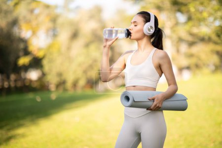 Téléchargez les photos : Une athlète sportive du millénaire caucasien vêtue de vêtements de sport élégants s'hydrate en buvant dans une bouteille d'eau, tenant un tapis de yoga, avec un casque dans un parc ensoleillé, en plein air - en image libre de droit