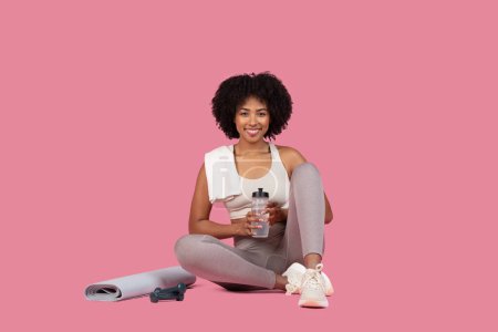 Foto de Feliz afroamericano fitness señora entusiasta toma descanso, sentado con botella de agua, esterilla de yoga, y saltar la cuerda, sonriendo a la cámara en el fondo rosa - Imagen libre de derechos