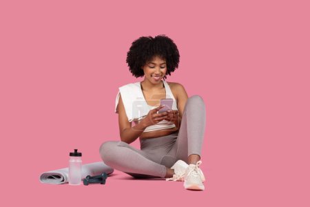 Téléchargez les photos : Jeune femme noire active prend une pause de la routine de remise en forme pour s'engager avec son smartphone, assis jambes croisées avec équipement de gymnastique sur fond rose - en image libre de droit
