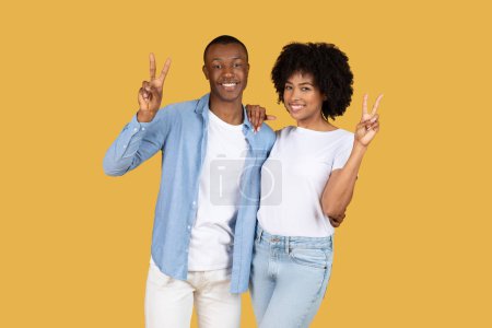 Téléchargez les photos : Un jeune couple noir joyeux portant des vêtements décontractés faisant des signes de paix sur un fond jaune moutarde solide avec espace de copie, studio. Relations, amour et mode de vie - en image libre de droit