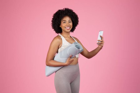 Foto de Mujer afroamericana joven radiante en traje de entrenamiento sonriendo para selfie, sosteniendo la esterilla de yoga bajo el brazo y usando el teléfono celular contra el vibrante fondo rosa - Imagen libre de derechos