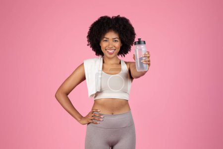 Photo for Radiant black fitness lady in workout gear proudly displaying her water bottle with white towel draped over one shoulder against pink backdrop - Royalty Free Image