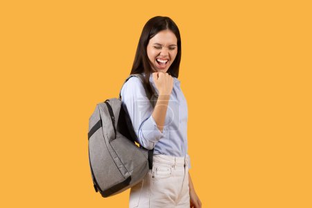 Photo for Cheerful young student wit backpack raises her fist in gesture of victory or excitement, embodying the feeling of success or win, possibly after receiving good grades in exams - Royalty Free Image
