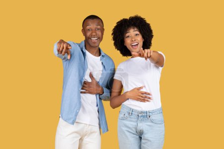 Photo for A joyful and laughing black young couple pointing at the camera, sharing a moment of fun and connection, set against a cheerful mustard yellow background, studio. Bullying, humor - Royalty Free Image