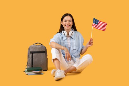 Téléchargez les photos : Étudiante optimiste assise avec le drapeau américain, exsudant un esprit patriotique et avide d'apprendre, avec son ordinateur portable et son casque suggérant une expérience éducative moderne et connectée - en image libre de droit