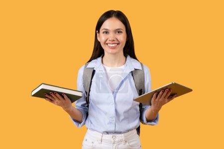 Lächelnde Studentin mit Buch in der einen Hand und modernem Tablet in der anderen, steht vor gelbem Hintergrund und symbolisiert die Schnittstelle zwischen klassischer und digitaler Bildung
