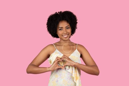 Téléchargez les photos : Radieuse jolie femme afro-américaine millénaire souriante avec une coiffure complète forme un cœur avec ses mains, mettant en valeur un geste d'amour sur un fond rose, studio - en image libre de droit