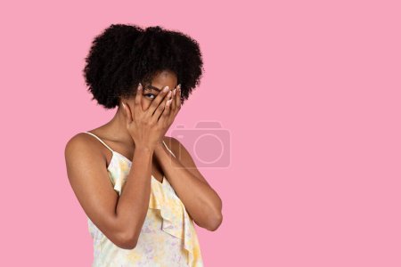 Photo for Curly-haired funny glad millennial African American woman playfully covers her face with hands, peeking through fingers, on pink background, studio. Fun, facial expression - Royalty Free Image