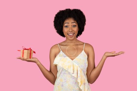 Foto de Sorprendida y encantada mujer afroamericana milenaria con el pelo rizado, sosteniendo un pequeño regalo con una cinta roja y haciendo gestos con su mano, sobre un fondo rosa, estudio - Imagen libre de derechos