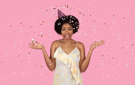 Téléchargez les photos : Femme afro-américaine millénaire extatique aux cheveux bouclés célébrant, portant un chapeau de fête et entouré de confettis, exprimant son bonheur avec un large sourire sur un fond rose - en image libre de droit