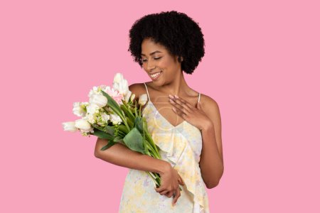 Photo for Content glad pretty millennial African American woman with a natural hairstyle admiring a beautiful bouquet of mixed flowers shes holding, on a soft pink background, studio - Royalty Free Image