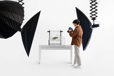 Téléchargez les photos : Dans un studio photo bien équipé, photographe masculin en tenue décontractée photographie méticuleusement des baskets vertes élégantes affichées sur une table blanche minimaliste - en image libre de droit