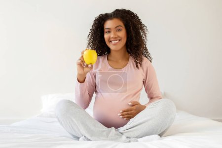 Foto de Feliz joven embarazada negra sosteniendo manzana mientras está sentada en la cama blanca, hermosa madre africana americana embarazada con ropa cómoda que exuda salud y felicidad en casa, sonriendo a la cámara - Imagen libre de derechos