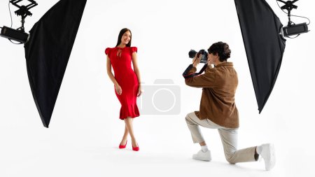 Téléchargez les photos : Scène studio de photographie dynamique avec photographe masculin concentré en tenue décontractée capturant la pose vibrante du modèle souriant en robe rouge magnifique - en image libre de droit