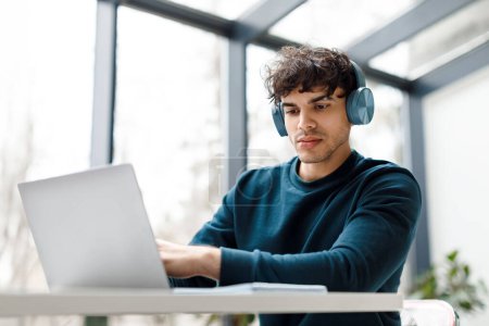 Konzentrierter junger europäischer Mann mit Kopfhörer sitzt am modernen Arbeitsplatz mit großem Fenster im Hintergrund und nutzt intensiv den Laptop