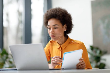 Konzentrierte junge schwarze Frau studiert intensiv an ihrem Laptop, während sie ihr Notizbuch in der Hand hält und in einem hellen, modernen Arbeitsraum mit natürlichem Licht sitzt