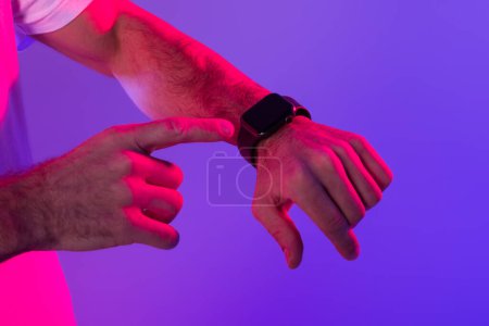 Téléchargez les photos : Homme méconnaissable pointant vers le tout nouveau gadget smartwatch avec écran noir vide sur son poignet, gros plan de la main, sur fond de studio éclairé de violet et bleu néon, maquette. Concept de dispositif technique - en image libre de droit