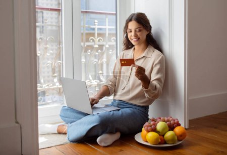 Foto de Feliz joven mujer cómodamente sentada en el suelo haciendo compra en línea con tarjeta de crédito y portátil, sonriente millennial compras femeninas en Internet, sentado junto al plato de frutas por la ventana en casa - Imagen libre de derechos