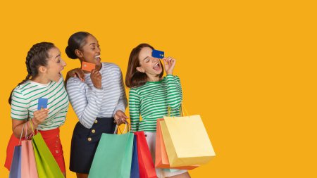 Photo for Cute young multiethnic women enjoying easy shopping, holding bank credit cards and colorful paper bags purchases, laughing, isolated on yellow studio background, panorama with copy space - Royalty Free Image