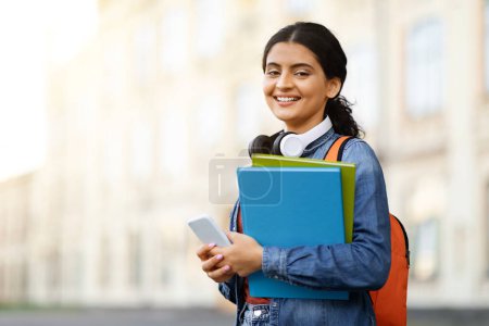 Photo for Portrait of cheerful young indian woman student with phone and exercise books in her hands posing outdoors next to university building, copy space. Educational mobile app - Royalty Free Image