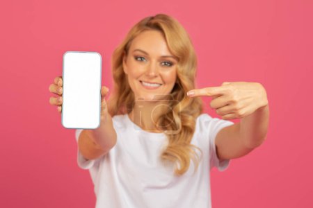 Photo for Smiling European blonde woman shows smartphone mockup pointing at empty gadget screen, showcasing communication or social media concepts, over pink studio background - Royalty Free Image
