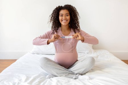 Photo for Overjoyed pregnant woman sitting cross-legged on bed and showing positive pregnancy test result, beautiful expectant mother beaming with happiness, expressing joy of upcoming motherhood - Royalty Free Image