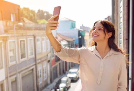Foto de Hermosa mujer joven tomando selfie con su teléfono inteligente en el balcón iluminado por el sol, feliz mujer milenaria disfrutando de la vibrante atmósfera de la ciudad, reflejando un estilo de vida urbano feliz y despreocupado, espacio libre - Imagen libre de derechos