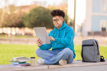 Brazilian college student guy websurfing on digital tablet, sitting with coffee and backpack, while learning online in park outdoor, immersed in educational website. E-learning platform offer