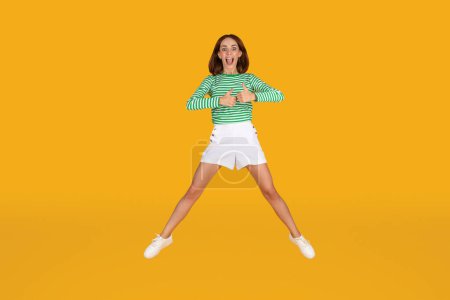 Photo for I like it. Positive pretty brunette young woman wearing summer outfit jumping over yellow studio background, grimacing and gesturing thumb ups, full length - Royalty Free Image