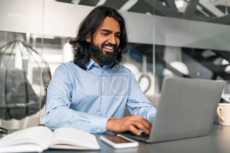 Photo for Handsome bearded long-haired millennial indian man wearing formal clothing manager sitting at desk at modern office, typing on laptop keyboard and smiling, sending emails, copy space - Royalty Free Image