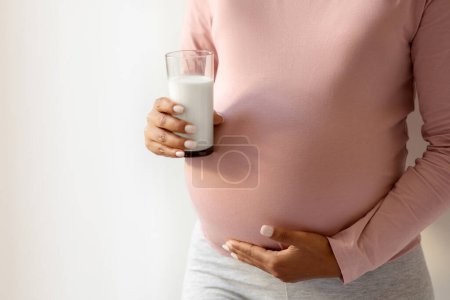 Téléchargez les photos : Femme enceinte berçant son ventre et tenant un verre de lait, femme enceinte noire méconnaissable signifiant l'importance de la consommation de calcium pendant la grossesse, plan recadré avec espace de copie - en image libre de droit