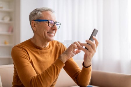 Photo for Grandfather wearing eyeglasses using cell phone at home. Happy elderly man sitting on couch in cozy living room interior, texting his family, scrolling, reading news online, copy space - Royalty Free Image