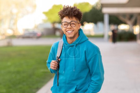 Photo for Cheerful brazilian student guy wearing eyewear and casual blue hoodie standing with his backpack and smiling to camera outside, on his way to college, posing in park at sunny day - Royalty Free Image