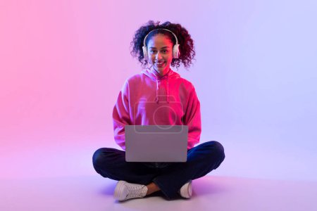 Téléchargez les photos : Contenu jeune femme portant un sweat à capuche rose, aime travailler sur son ordinateur portable tout en portant un casque, assis jambes croisées sur le sol contre fond dégradé coloré néon - en image libre de droit