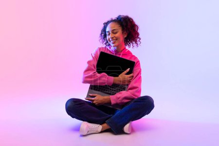 Foto de Encantada dama negra con el pelo rizado, abraza su portátil, sentado con las piernas cruzadas en rosa degradado a fondo azul, exudando felicidad y satisfacción - Imagen libre de derechos