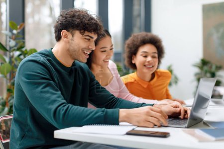Drei fröhliche Studenten unterschiedlicher ethnischer Herkunft nehmen an einer gemeinsamen Lerneinheit mit Laptop teil, umgeben von üppigen Zimmerpflanzen