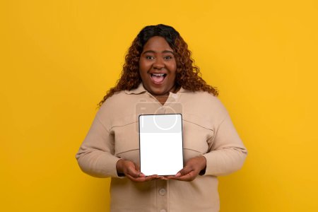Foto de Mujer americana africana millennial gordita positiva mostrando tableta digital con pantalla blanca en blanco, mostrando una buena aplicación móvil o recomendando oferta en línea, fondo amarillo, maqueta, espacio de copia - Imagen libre de derechos