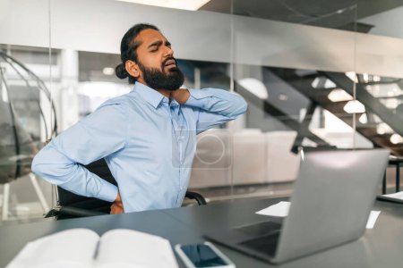 Photo for Indian man feeling neck and back pain, suffering from discomfort and stress while working on laptop at modern office setting, tired eastern guy sitting at desk, massaging painful area, copy space - Royalty Free Image