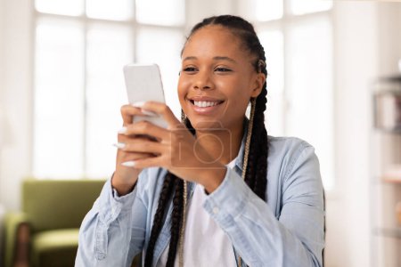 Téléchargez les photos : Joyeuse jeune femme noire avec des tresses souriantes tout en s'engageant avec son smartphone, peut-être pendant la pause des études en ligne ou en profitant des médias sociaux - en image libre de droit