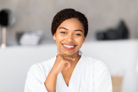 Photo for Radiant young black woman with beautiful skin smiling at camera, happy african american female wearing white bathrobe posing in bathroom interior at home, enjoying self-care routine, copy space - Royalty Free Image