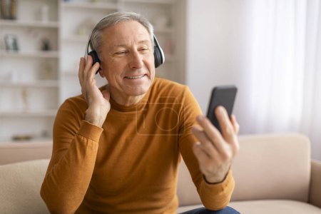 Foto de Hombre europeo de pelo gris positivo sentado en el sofá en casa, usando auriculares inalámbricos y teléfono celular, viendo contenido de video, escuchando música, espacio para copiar - Imagen libre de derechos