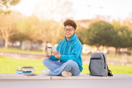 Téléchargez les photos : Étudiant universitaire brésilien gars profiter du café et en utilisant son smartphone pour étudier en ligne, prend une pause dans le parc en plein air, immergé dans les loisirs de l'éducation moderne, souriant à la caméra - en image libre de droit