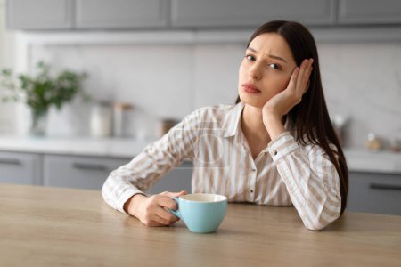 Photo for Pensive sad young woman holding blue coffee cup, resting her cheek on her hand, sitting at wooden kitchen table with concerned expression, free space - Royalty Free Image