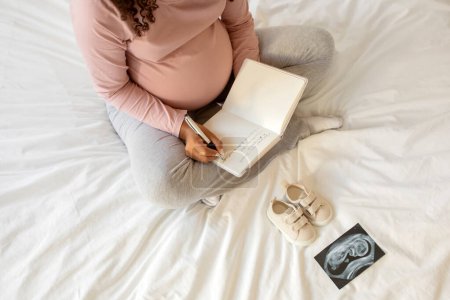 Téléchargez les photos : Femme enceinte écrivant des pensées dans son carnet de notes à la maison, assise sur le lit entourée de chaussures minuscules et d'une image d'échographie, future mère créant des souvenirs pendant sa grossesse ou faisant une liste de contrôle - en image libre de droit