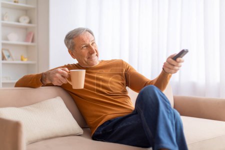 Téléchargez les photos : Joyeux beau senior allongé sur le canapé à la maison, tenant la télécommande, buvant du café et changeant de chaînes sur la télévision. Grand-père regarder des films ou des nouvelles du matin, copier l'espace - en image libre de droit