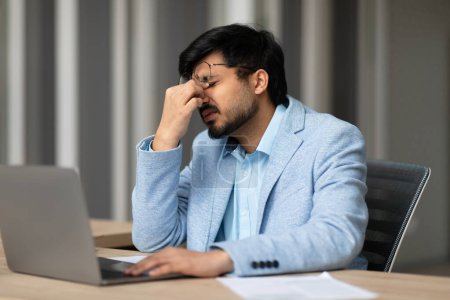 Indian businessman experiencing eye strain and fatigue sitting tired after computer work in office interior. Headache and health problems during stressful day at workplace