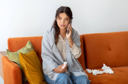 Photo for Concerned young woman wrapped in blanket making phone call to doctor, ill millennial female sitting on couch with used tissues scattered around, feeling unwell at home, suffering seasonal cold - Royalty Free Image